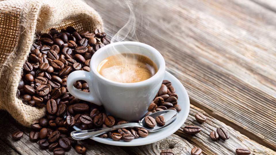 Saco con granos café tostados que caen junto a un pocillo blanco con café caliente
